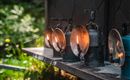 A row of old oil lamps stands on a shelf. In the background, green plants are visible, creating a natural environment.