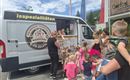 An ice cream truck with various ice cream specialties is parked on the street. Children and adults are standing in line to buy ice cream.