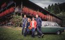 Eine Gruppe von vier Personen steht vor einem Holzhaus mit blühenden Balkonpflanzen. Ein grünes Auto befindet sich neben ihnen auf einer Wiese.