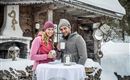 Ein Paar steht lächelnd an einem Tisch im Schnee und hält Tassen in der Hand. Im Hintergrund ist eine rustikale Hütte und gestapeltes Holz zu sehen.