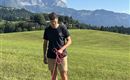 A man is standing on a meadow holding two Corgis on leashes. In the background, mountains and a blue sky can be seen.