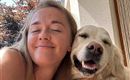 A woman smiles happily with a dog beside her.  
They enjoy a sunny day outdoors.