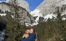 A woman holds a dog in her arms and smiles in nature. In the background, tall mountains and a clear blue sky can be seen.