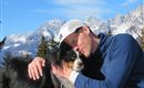A man hugs his dog in the mountains. In the background, snow-covered mountains and a blue sky can be seen.