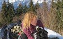 A person with long hair is sitting in the snow and petting their dog. In the background, impressive mountains and a blue sky can be seen.