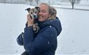 A smiling woman is holding a small dog in her arms. In the background, there is snow and it is lightly snowing.