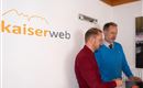 Two men are standing at a table and looking at a laptop. In the background, the logo of kaiserweb can be seen.