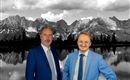 Two men in suits are standing in front of an impressive mountain landscape. The background shows the mountains and a calm lake, while the sky is cloudy.
