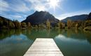 A quiet lake with a wooden pier and colorful trees in the background. The sun shines in the clear sky and reflects in the water.