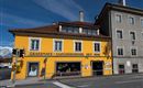Ein gelbes Gebäude mit dem Schriftzug "Grassmayr - Glockengießerei seit 1599" steht an einer Straßenecke. Daneben befindet sich ein graues Haus und im Hintergrund sind Berge zu sehen.