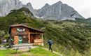 Eine Berghütte inmitten von grünen Wiesen und hohen Bergen. Ein Wanderer nähert sich der Hütte.