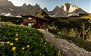 Eine gemütliche Hütte steht vor beeindruckenden Bergen. Der Weg zur Hütte ist von grünem Gras und bunten Blumen umgeben.