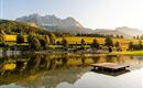 Ein malerischer See mit sanften Hügeln und reflektierenden Bergen im Hintergrund. Die goldene Abendsonne beleuchtet die Landschaft und schafft eine ruhige Atmosphäre.