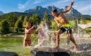 Eine Gruppe von Kindern spielt im Wasser und splasht herum. Im Hintergrund sind Berge und grüne Bäume zu sehen.