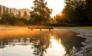 Ein ruhiger See im Morgenlicht mit einer sanften Dunstschicht. Im Hintergrund sind Bäume und die aufgehende Sonne zu sehen.