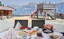 A set table with breakfast outdoors, including pastries, jam, and coffee. In the background, a playground with a slide and the mountains can be seen.
