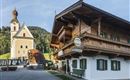 A traditional old inn with wooden decoration and green shutters stands next to a beautiful church. In the background, green hills and a clear sky are visible.