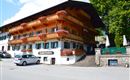 A traditional wooden house with balconies decorated with flowers. Additionally, a green surrounding and a parking lot in front.