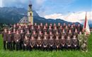Eine Gruppe von Männern und Frauen in Uniform steht in einer grünen Wiese vor einer Kirche und Bergen im Hintergrund. Sie posieren gemeinsam für ein Gruppenfoto.