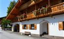 A traditional chalet with wooden balustrades and flower boxes. The street in front of the building is quiet and lined with trees.