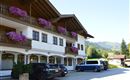 A picturesque building in the Bavarian style with balcony flowers. In the foreground, several cars are parked in front of a beautiful mountain landscape.