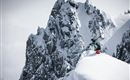 Ein Skifahrer steht auf einem schneebedeckten Gipfel mit beeindruckenden Felsen im Hintergrund. Die Szene vermittelt ein Gefühl von Abenteuer in einer winterlichen Berglandschaft.