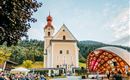 A picturesque church with a red tower stands in the background. In front of the church, a concert is taking place in a wooden pavilion, surrounded by an audience and green nature.