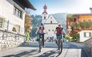 Two women are riding their bicycles through a picturesque village. In the background, there is a church, and the surroundings are bordered by mountains.