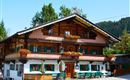 A traditional wooden house in alpine style with green shutters and blooming balconies. It is located surrounded by trees and a clear blue sky.