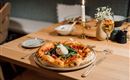 Een tafel met een heerlijke pizza, belegd met verse groenten en een ei. Daarnaast staat een glas champagne en een kaars zorgt voor een gezellige sfeer.