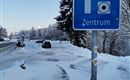 Ein Parkplatzschild mit der Aufschrift "Zentrum" steht in einer verschneiten Umgebung. Im Hintergrund sind schneebedeckte Bäume und ein Weg zu sehen.