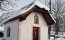 Eine kleine Kapelle mit einem Schnee bedeckten Dach. Umgeben von winterlicher Landschaft und schneebedeckten Bäumen.