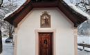 Eine kleine Kapelle mit einem schneebedeckten Dach steht in einer winterlichen Landschaft. Umgeben von schneebedeckten Bäumen und einem ruhigen, friedlichen Ambiente.