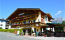 A charming alpine hotel with wooden decor and balconies. In the background, beautiful mountains and a clear blue sky can be seen.