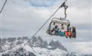 Eine Skiliftkabine mit zwei Fahrgästen schwebt über schneebedeckte Berge. Der Himmel ist bewölkt und die Landschaft wirkt winterlich.