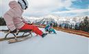 Zwei Kinder rodeln auf einem verschneiten Hang. Im Hintergrund sind schneebedeckte Berge und ein klarer Himmel zu sehen.