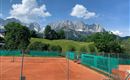 Ein Tennisplatz umgeben von grünen Bäumen und majestätischen Bergen im Hintergrund. Der Himmel ist klar und blau mit einigen weißen Wolken.