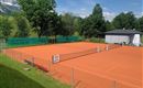 Ein Tennisplatz mit roter Asche, umgeben von Bäumen und einem klaren blauen Himmel. Im Hintergrund steht ein kleines Gebäude.