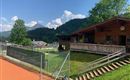 Ein sportlicher Bereich mit einem Tennisplatz und einem Holzhaus im Hintergrund. Die Umgebung ist grün, umgeben von Hügeln und Bäumen, und der Himmel ist blau mit einigen Wolken.