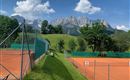 Ein Tennisplatz mit rotem Sand und rundherum grünen Wiesen. Im Hintergrund sind beeindruckende Berge und ein blauer Himmel mit einigen Wolken zu sehen.