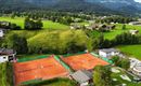 Eine Tennisanlage mit zwei Plätzen auf rotem Sand in einer grünen Landschaft. Im Hintergrund sind Berge und Wohnhäuser zu sehen.
