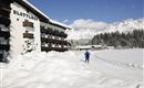 Ein Hotelgebäude mit dem Namen "Blatthof" liegt in einer schneebedeckten Landschaft. Im Hintergrund sind schneebedeckte Berge und ein klarer blauer Himmel zu sehen.