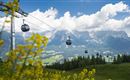 A landscape with mountain gondolas traveling over green meadows. In the background, impressive mountains and a blue sky with clouds can be seen.