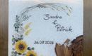 Eine dekorative Tafel mit der Aufschrift "Sandra & Patrick" und dem Datum "26.09.2026". Umgeben von Blumenmotiven und einer Holzstruktur, die eine warme Atmosphäre schafft.