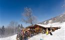 Eine alpine Skihütte steht im Schnee unter einem blauen Himmel. Skifahrer genießen die winterliche Landschaft und machen eine Pause.