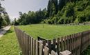 A large, green field surrounded by a wooden fence. In the background, trees and a clear sky can be seen.