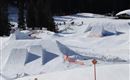 Eine verschneite Landschaft mit mehreren Sprüngen für Wintersportler. Im Hintergrund sind Bäume und ein Gebäude sichtbar.