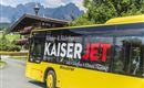 A yellow bus with the inscription "KaiserJET" drives through a picturesque landscape. In the background, mountains and a cozy alpine hut can be seen.