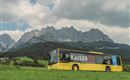 A yellow painted bus is standing on a green meadow, surrounded by impressive mountains. The sky is partly cloudy, creating a picturesque landscape.