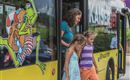 A woman and two girls are getting out of a colorful bus. The bus has a striking design featuring a drawing of a frog character.